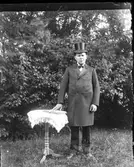 En man iklädd rock och cylinderhatt står i en trädgård vid ett litet pelarbord med duk och håller handen på en uppslagen bok, som ligger på bordet.