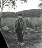 En man står barfota utomhus i gräset med åkerlandskap och berg i bakgrunden. (Negativet är skadat i vänster nedre hörn)
(Se även bild GEA075 och GEA077)