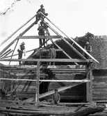 Några män bygger stommen till en tillbyggnad av en gammal ladugård. En man sitter uppe på takåsen och under honom står en man med såg på stege. Övriga tre håller också i olika verktyg som yxor och sågar. Nedtill sitter en pojke i kubb. Den äldre byggnaden har stomme av skiftesverk och halmtak, medan tillbyggnadens stomme blir av resvirke.