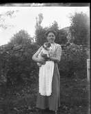 En ung kvinna i spetsförkläde står utomhus med en liten hund på armen. (Se även bild nr GEB248)