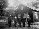 Fem män står vid en husgavel, där två ingångar med spegeldörrar finns. Tre av männen har blommor fästa vid kavajslagen och tre har fickur med kedjan fäst i västen. På husnocken sitter en flaggstång och bakom stugan ligger en stengärdsgård. (Bilden beskuren pga skador)