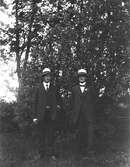 Två yngre män står finklädda utomhus med likadana ljusa, toppiga hattar med mörka band och den ene bär en blomma på kavajslaget.