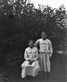 Två flickor, troligen systrar, i likadana klänningar med sjömanskragar. De står framför ett lönnbuskage.