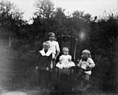Fyra finklädda barn är samlade för fotografering utomhus. Den minsta sitter i en gungstol draperad med en vävd textil och gossen till höger sitter på en gunghäst på hjul.