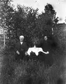 Ett äldre par sitter i en trädgård vid ett bord med duk. Han har skepparkrans och en klockkedja i västen. Hon bär huckle och mörk dräkt med en brosch i halsen, håller en bok i knät och med andra handen håller hon om vasen på bordet.