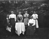 Ett sällskap om sju personer i olika åldrar fotograferade utomhus vid foten av ett berg. De äldsta sitter vid ett bord och kanske är de fyra kvinnorna deras döttrar. En lite pojke står hos mannen, som håller i en Norra Hallands Tidning Vestkusten. De äldsta är mycket solbrända - mannens panna är vit jämfört med ansiktet i övrigt.