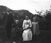 En familj med tre barn fotograferade utomhus med stengärdsgård och kala berg i bakgrunden. Modern sitter med minsta barnet i knät och bär en smalrantid klänning med klädda knappar. Pojken har på sig en kolt och flickan till höger en skir spetskrage på klänningen med stråveck och klädda knappar. (Se bild på endast henne GEB187)