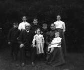 Familj med sju barn samlade utomhus för fotografering. Föräldrarna sitter och barnen står runtom. Tre pojkar har likadana jackor med rundade kragar. (Se bild på de två minsta barnen GEB184)