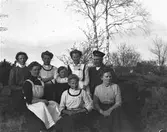 Sju flickor och en man, Sällstorpsbor, sitter på en bergknalle och de flesta bär en Majblomma, så det lär vara kring 1:a maj. Två har likadana klänningar och broscher och två andra bär förkläden; möjligen är detta en grupp med tjänstefolk av olika slag, förutom flickan.
(Bilden beskuren pga skador på negativet)