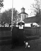 Två hattklädda unga kvinnor står utanför kyrkan i Karl Gustav med var sin liten hund. Bakom dem står grinden öppen i trästaketet som hägnar en trädgård med flaggstång. (Kvinnan till höger finns även på bild nr GEB166)