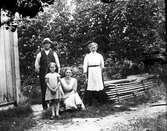 En familj med två döttrar samlad vid husknuten, eventuellt Hornagård i Sällstorp. Till höger ligger brädor på tork.