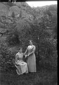 Två unga kvinnor utomhus med ett berg i bakgrunden. Den ena sitter på en pinnstol och den andra står bredvid. Stolen och dahliorna till vänster tyder på att de är i en trädgård som gränsar till berget.