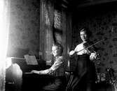 Två unga kvinnor musicerar i ett rum med stora vallmoblommor på tapeten. Den ena spelar tramporgel till noter och den andra spelar fiol med blicken på fotografen.