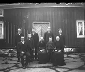 Familjen samlad på den stenlagda gården framför manhuset på Ulvatorp 1, Kullen i Sällstorp. Alla är mörkt klädda, i mitten sitter en äldre kvinna omgiven av sju stående pojkar och sannolikt deras föräldrar som sitter. Männen bär alla fluga, utom den minsta pojken som har sjömanskrage. Kvinnorna har örhängen och broscher; den äldre kvinnans brosch påminner om ett dräktsmycke.