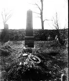 Grav vid obeliskformad gravsten för Lovisa Svensson, död 30 januari 1909. Gravkullen täcks av granris, en blomsterbukett och två s k pärlkransar. Detta blev från omkring sekeskiftet 1900 en vanlig gravdekoration i södra Sverige. Modet spreds från Frankrike på 1870-talet och tillverkningen kom till stor del att ske i södra Tyskland. Pärlkransarna bestod av sammanflätad ståltråd som dekorerades med glaspärlor i olika färger och modeller, vilka börjat massproduceras. I mitten av kransen lades en religiös figur skapad av gips, celluloid eller annat, t ex ett kors eller en ängel, under en glaskupa. Text kunde också införlivas i dekoren. Pärlkransarna importerades och kunde köpas av pappershandlar, handelsträdgårdar m fl. På somliga ställen kunde fattiga låna en pärlekrans.