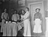 Fem kvinnor står vid ett runt bord i en salong och stryker löskragar. De bär förkläden så troligen är de tjänstefolk. På bordets tjocka duk har de lagt ett lakan och därpå en strykbräda. Två av kvinnorna har strykjärn medan en har händerna i ett lerfat.