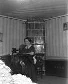 En kvinna sitter vid en trampsymaskin intill en äldre, plan kakelugn på träben med melerat kakel. På kistan vid väggen står initialerna BGYD och årtalet 1877. Väggen har en slät träpanel nedtill och däröver randiga tapeter.