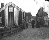 Ett äldre par står med en liten pojke och en äldre flicka utanför sin gård. Mellan bostadshuset till vänster och en ekonomibyggnad till höger är ett plank uppfört som hindrar infart och insyn till gårdsplanen. På ena gaveln har vissa bräder och lister nyligen bytts ut.
(Negativet skadat till höger)