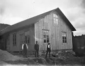 Tre snickare poserar med sina verktyg framför en stor tillbyggnad till ett äldre hus. Byggnaden har profilerad locklist, står på stengrund och har stora spröjsade fönster med korspost. Längs grunden ligger en lång stege och männen har såg, hammare, yxa och slägga till hands.