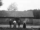 Ett äldre par och troligen deras dotter och hennes son står framför en stuga med åker i bakgrunden. Längs med stugan går byvägen. Huset är av typen parstuga, dvs med en centrerad ingång och ett rum på var sida. Taket har haft halmtäckning (syns på skorstenen) men numera täcks det av stickor (pärt).