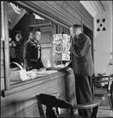 En man står och pratar med en kvinna i receptionen på Abisko turiststation. Intill dem finns ett ställ med vykort. Interiör.