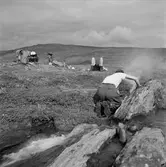 Dag Hammarskjöld på fjällvandring på Sulitelma. Två män har slagit läger, den ene ligger på rygg och vilar medan den andre gör upp eld vid ett klippblock.