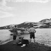 Dag Hammarskjöld och Sverer Åström på fjällvandring på Sulitelma. Två män står på stranden intill packning och utrustning. De ser ut över vattnet där ett sjöflygplan landat.