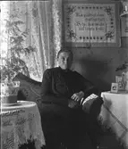 Småskollärarinnan Bertha Engström i Sällstorp sitter i lärarbostadens finrum. Till den mörka klänningen bär hon brosch, långt halsband och ringar. Krukan till rumsgranen på bordet har någon sorts pappersomslag.