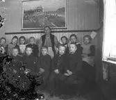 Småskollärarinnan Bertha Engström i Sällstorp tillsammans med sin klass inne i skolan. Väggen är spontad och avdelas med en smal list. Upptill hänger en plansch som synes avbilda olika moment inom jordbruket. (Negativet skadat i vänster sida)