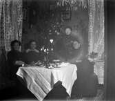Bjudning i juletid hos småskollärarinnan Bertha Engström i Sällstorp. Bordet är dukat vid julgranen och förutom Bertha till höger ses tre kvinnliga gäster. Kakor, bullar och uppskuren citrusfrukt, troligen apelsin, ligger på olika fat. Några koppar eller dryck finns emellertid inte på bordet, som pryds med en hög kulsvarvad träljusstake med glasmanschetter.