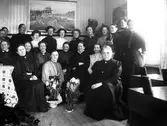 En grupp kvinnor och en man samlade i småskolan i Sällstorp. Kanske firas någon eller något då det står krukor med hyacinter och narcisser på golvet. Kvinnan som sitter närmast blommorna är Bertha Engström som undervisar i skolan. Hennes systrar sitter längst fram, Constance Engström till höger, som är folkskollärare i socknen, och Fredrika Engström till vänster. (Se även bild nr GEB273)