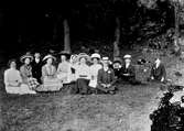 En grupp ungdomar sitter i en skogsdunge nedanför ett berg med en kaffebricka mitt i bland sig. Till höger sitter tre gossar och fotografen själv i ljus hatt, Gunnar Engström. Damerna har breda sommarhattar och möjligen är det midsommar eftersom kvinnan i mitten bär folkdräkt.