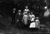 En familj fotograferad utomhus; ett par med fyra barn samt en äldre man till vänster. (De två små flickorna i rutiga klänningar finns även på bild GEB212 och mannen till vänster på GEB118)
