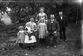 En familj fotograferad utomhus under aspträd; ett par med fyra barn samt en äldre man till höger. Till vänster om dem och framför modern ligger ett krossat porslinsföremål och mellan träden i bakgrunden hänger linor. 
(Se även bilderna GEA075, GEB212 och GEB118)