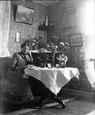 En ung kvinna i smårutig klänning sitter vid ett pelarbord i salongen. Förmodligen är det Alma Börjesson (gift Andersson, se bildnr GEB119). Hon förefaller ha en bemärkelsedag eftersom hon håller på att läsa igenom en hög med post framför sig och såväl bord som skänk smyckas av blombuketter och krukväxter, prydda med kräppat papper.