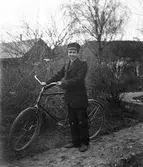 En yngling står i en trädgård på en kantskuren grusgång med en cykel och håller hårt i styret. I bakgrunden ses gårdsbyggnader.