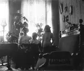 Interiör med en kvinna och två små barn i motljus från salongens stora fönster. Rummet har högt till tak och randiga tapeter med stora blommor som går samman med gardinernas rosenmöster, krukväxter och blombuketter som pryder rummet. Möblerna bär sen 1800-talsprägel och på bordet står en hög, smal fotogenlampa.