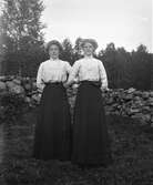 Två unga kvinnor, systrar sannolikt, står i armkrok och likadana kläder framför en stengärdsgård. De har höghalsade vida blusar med krås och skärp med rosett i midjan till långa mörka kjolar. (Se även bildnr GEB195)