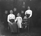 Sju syskon samlade utomhus där äldst och yngst i skaran sitter på stolar. De tre äldsta bröderna bär likadana storkragade rockar och den lille en kolt. (Se även bildnr GEB184, GEB201)