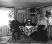 Syllinge, Jonasa i Veddige. Jenny och Johan sitter i finrummet vid bordet, där det ligger en kraftigt mönstrad duk. Soffan pryds av virkade antemarkasser och ovanpå trasmattorna ligger ytterligare en matta under bordet och stolarna där paret sitter. Bakom dem står piedestaler med krukväxter. Randiga jugendtapeter klär väggarna och av bonaden att döma är bilden spegelvänd.
(Se även bildnr GEB198)