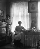 Två kvinnor, troligen mor och dotter, i motljus vid stort fönster. Dottern står stödd mot en sekretär och modern sitter vid ett bord med en virkad duk med mycket långa fransar.