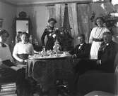 Sex ungdomar samlade kring ett tebord, två män och fyra kvinnor, varav en håller i en bok. På bordet står en teservis av metall och en rosenbukett. På väggen klättrar en murgröna. (Se även bildnr GEB207)