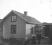 Ett par står utanför sitt hus en solig dag. Mannen bär kostym och kubb, kvinnan en storrutig blus till den hellånga mörka kjolen. Huset är målat, har en liten glasveranda, fönster med korspost och diskreta figursågade kanter längs takfoten på både hus och veranda. I bakgrunden ses ett uthus målat i mörkare färg. (Möjligen samma par på bildnr GEB276)