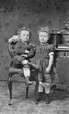 Ateljéporträtt av de två små syskonen Emma och Hjalmar Svensson. Hjalmar sitter på en stol i vuxen position med både armar och ben i kors. Emma står vid en byrå med ena handen på lillebroderns axel. Båda har snörkängor på sig.