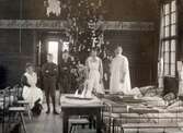 Patienter och sjuksystrar i julpyntad sjuksal i Kustsanatoriets träpaviljong från 1904. En rikt pyntad och hög gran står i centrum, en lång bonad pryder väggenarna och på bordet står en julbukett med tulpaner.