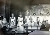 Matbespisning till små patienter på Kustsanatoriet Apelviken i träpaviljongen från 1904. Här vårdades skrofulösa (tbc) barn och ungdomar sommartid. På väggen bakom kökspersonalen hänger en stor tavla med sommarmotiv till prydnad.