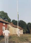 Staffan Bjerrhede halar ner flaggan efter hembygdsföreningens midsommarfirande 1993. Berny Gustafsson och okänd man ser på. Till vänster ses den röda Hembygdsgården Långåker 1:3 och i bakgrunden Långåker 1:2 