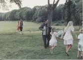 Midsommarfirande vid Hembygdsgården Långåker 1:3 1993. 
Till vänster ses folkdräktsklädde Staffan Bjerrhede promenera. Barn dansar kring midsommarstången till dragspelsmusik av okänd musikant från Göteborg.
Relaterade motiv: A03389 - A03408.