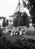 Ringlekar runt midsommarstången på Kustsanatoriet Apelviken under 1950-talet. Till vänster om stången sitter musikerna; en kvinna med dragspel och en man med gitarr. Kanske spelas 