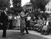 Kung Gustav VI Adolf på Eriksgata genom Halland, då han besökte Kustsanatoriet Apelviken i Varberg. Till vänster om kungen går Harald Liljenberg, intendent vid sanatoriet, och överläkare Albert Ahlberg går bredvik majestätet, som skymmer honom. Här hälsar kungen på en rad kvinnliga patienter.